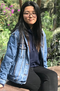 Student in denim jacket and glasses sitting outside by trees and plants